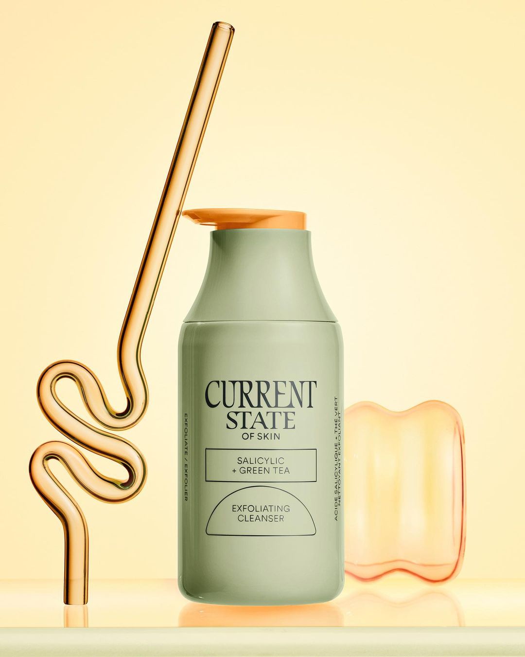 Bottle of exfoliating cleanser with a glass straw and orange facial massage tool on a shiny surface against a yellow background.