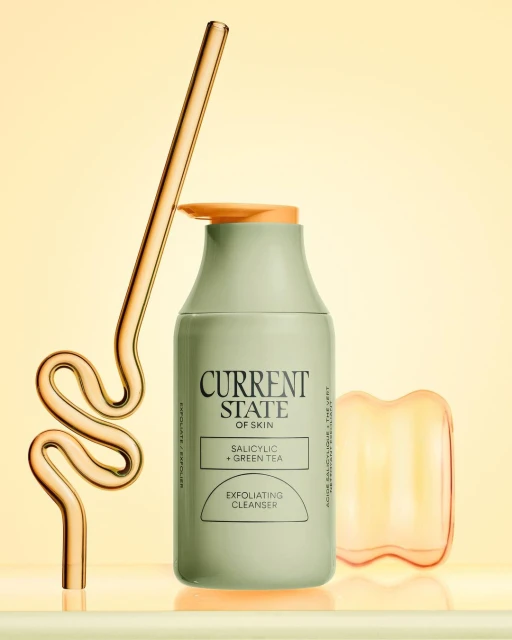 Bottle of exfoliating cleanser with a glass straw and orange facial massage tool on a shiny surface against a yellow background.