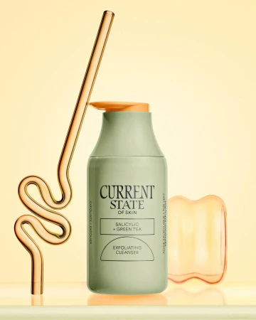 Bottle of exfoliating cleanser with a glass straw and orange facial massage tool on a shiny surface against a yellow background.