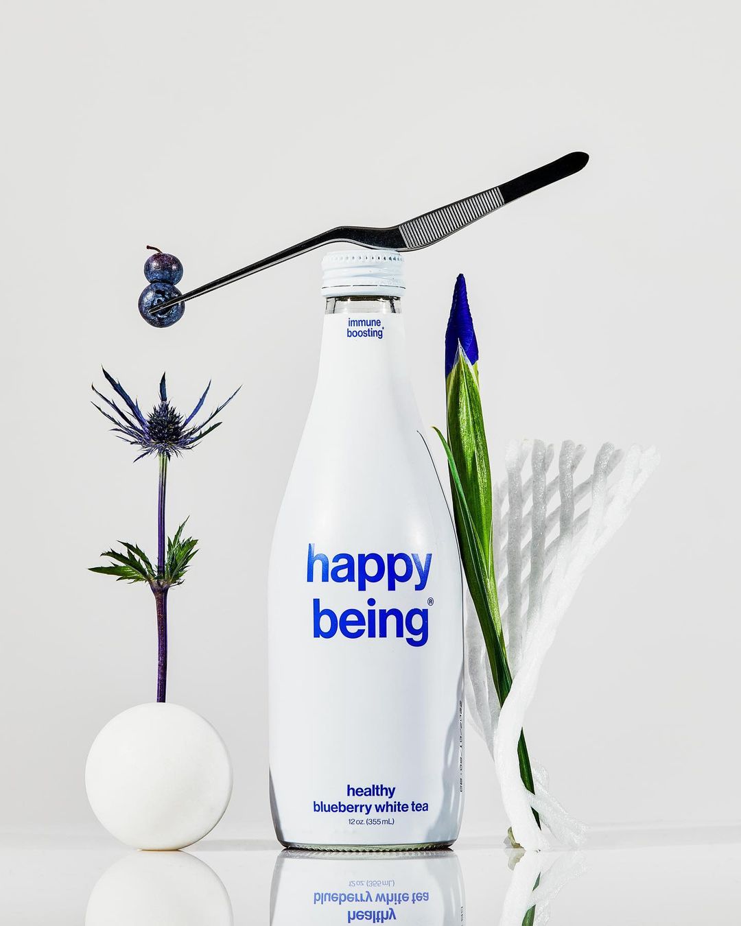 Still life of blueberries on a metal tool over a white drink bottle, accompanied by thistle, iris, and white paper.