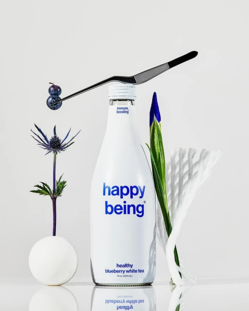 Still life of blueberries on a metal tool over a white drink bottle, accompanied by thistle, iris, and white paper.