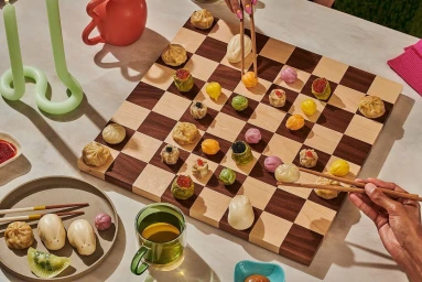 Overhead shot of a wooden chessboard used as a game board with dumplings replacing chess pieces, played by hands with chopsticks.