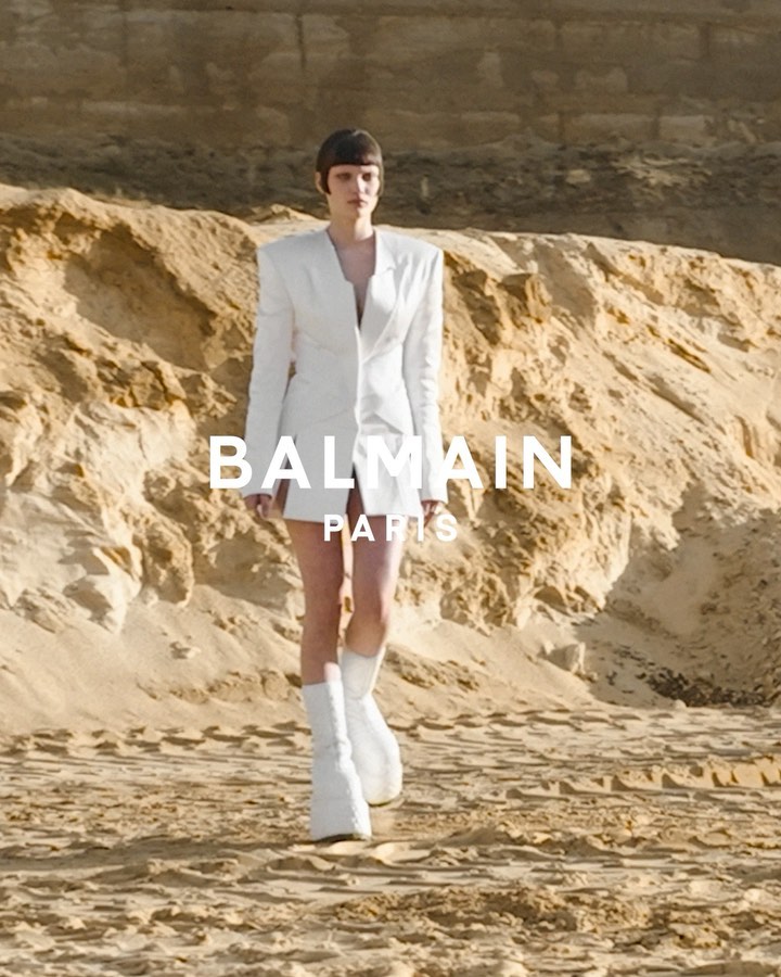 A woman models a white blazer dress and white boots on a tan sandy terrain, in a fashion editorial with the Balmain logo visible.