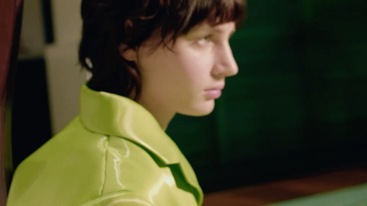 Profile of a young adult in a neon yellow jacket, looking away in an indoor studio, with blurred dark-green and red background.