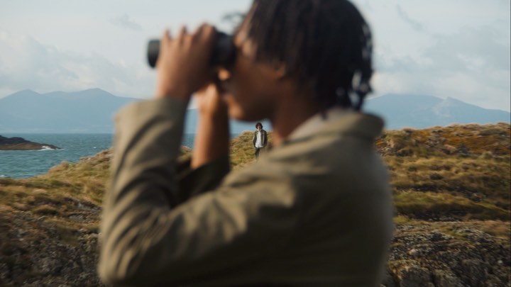Man in a green jacket uses binoculars on a grassy hilltop overlooking the ocean and mountains, while another man stands further away.