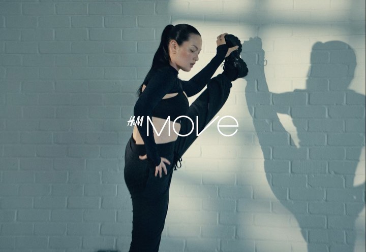 Athletic woman in black activewear showcasing flexibility in a dynamic pose against a grey brick wall. H&M Move branding is visible.