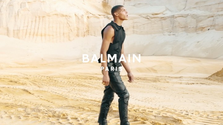 A male fashion model poses in black tactical vest and leather pants against a sandy desert backdrop, showcasing an edgy editorial style.