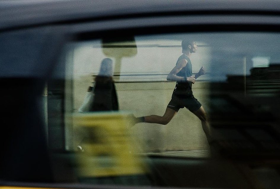 A man in athletic wear is running outside; a car window's reflection shows a blurred figure in the reverse direction.