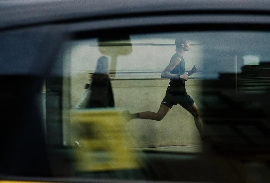 A man in athletic wear is running outside; a car window's reflection shows a blurred figure in the reverse direction.