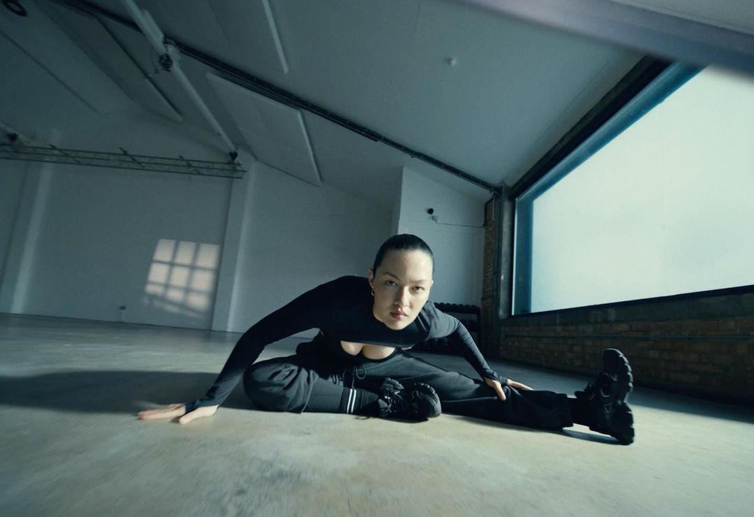 Asian adult woman in a black long-sleeved shirt and black sweatpants sitting on a concrete floor stretching her legs in a sparse studio.