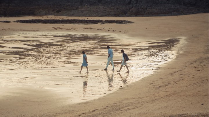 Three people walk along a sandy beach at low tide with a rocky outcrop in the background, with their reflections visible in the wet sand.