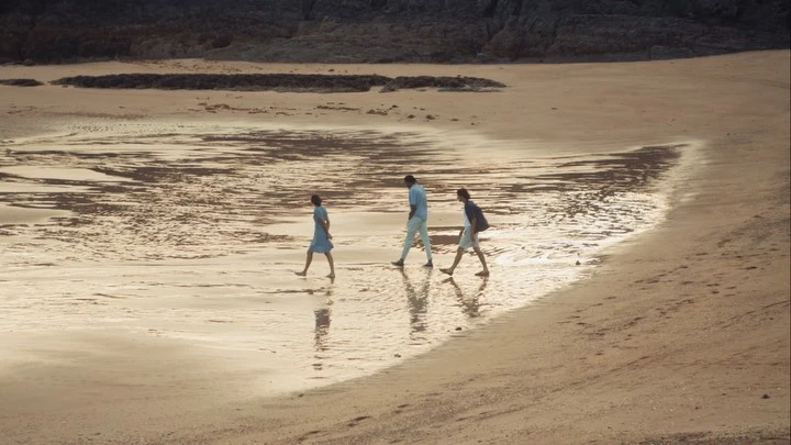 Three people walk along a sandy beach at low tide with a rocky outcrop in the background, with their reflections visible in the wet sand.
