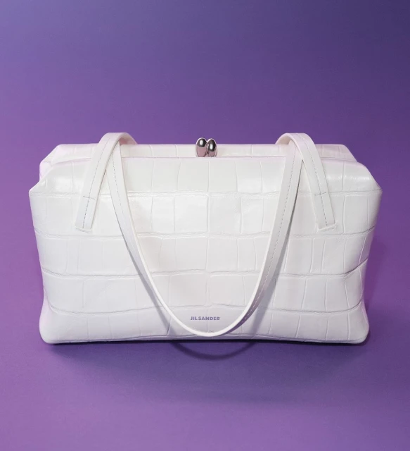 White Jil Sander handbag with a faux crocodile texture and silver hardware against a solid purple background in a studio shot.