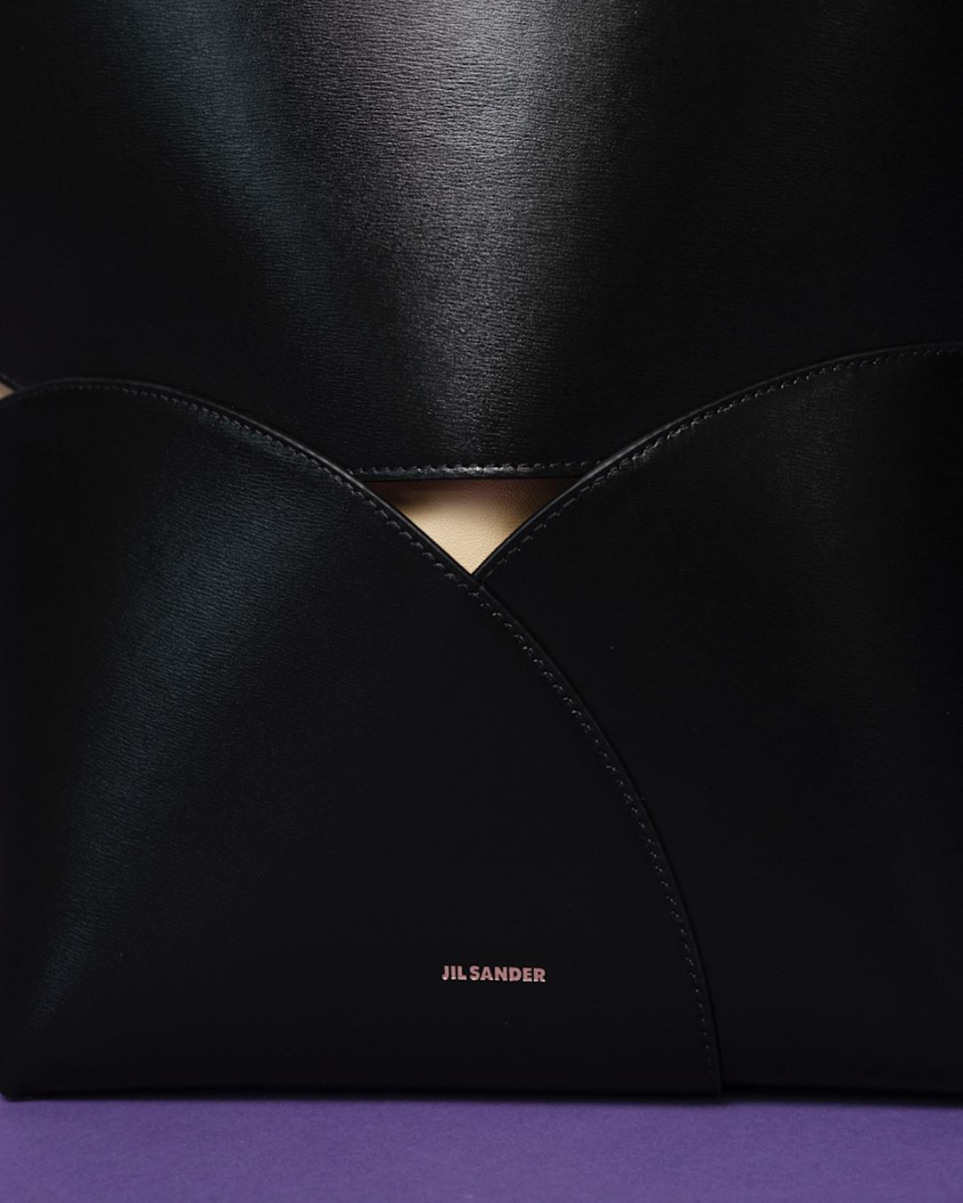 Close-up of a black leather Jil Sander tote bag with a modern design, featuring a geometric flap and a purple backdrop.