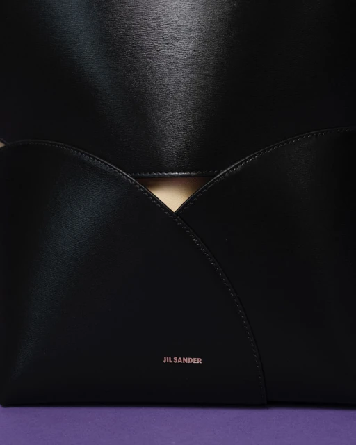 Close-up of a black leather Jil Sander tote bag with a modern design, featuring a geometric flap and a purple backdrop.