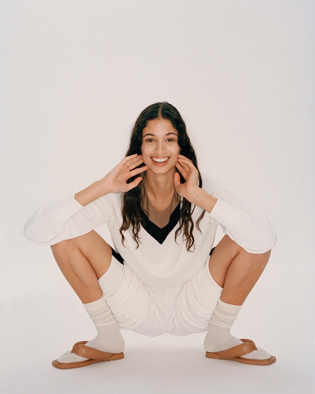 Smiling woman in a white sweater and shorts is squatting down with her hands near her face in front of a white background.