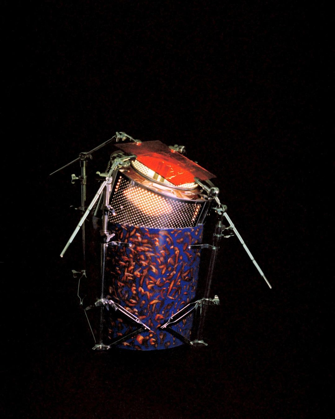 Unique drum-like percussion instrument constructed with metal supports, orange membrane, blue patterned body, illuminated from inside in darkness.