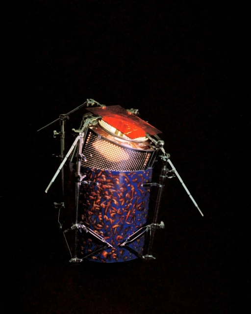 Unique drum-like percussion instrument constructed with metal supports, orange membrane, blue patterned body, illuminated from inside in darkness.