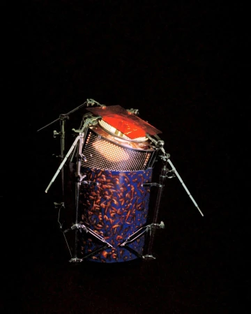 Unique drum-like percussion instrument constructed with metal supports, orange membrane, blue patterned body, illuminated from inside in darkness.