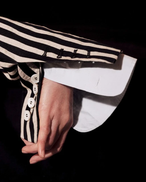 Close-up of a person's hand intertwined, wearing a striped blazer over a white shirt against a dark background. Focus on texture and detail.