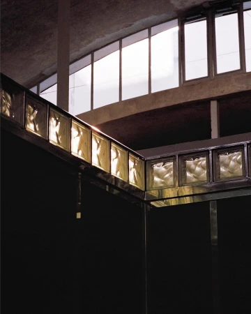 Architectural interior featuring light filtering through rectangular glass blocks set in a metal frame with high windows and a dark backdrop.