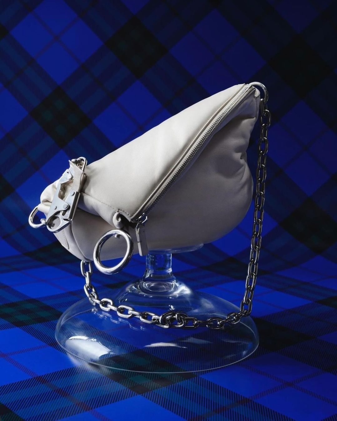 Ivory-colored, pillow-shaped handbag with a silver chain and hardware, displayed on a transparent glass goblet against a blue tartan backdrop.