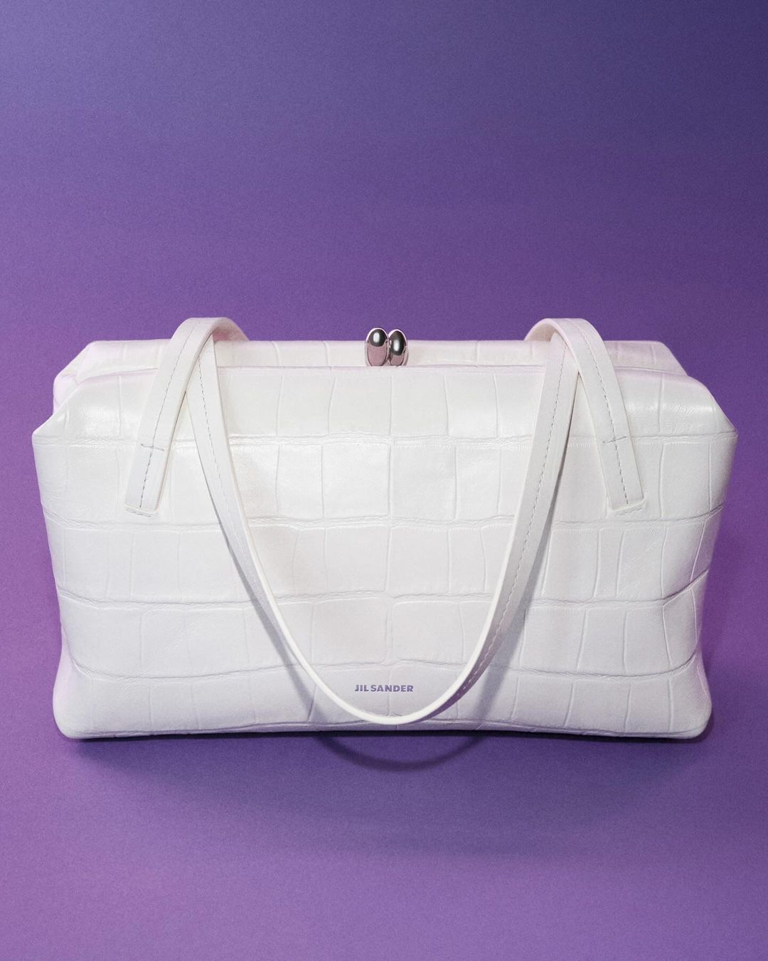 Studio shot of a white Jil Sander handbag with a crocodile print set against a solid purple backdrop.
