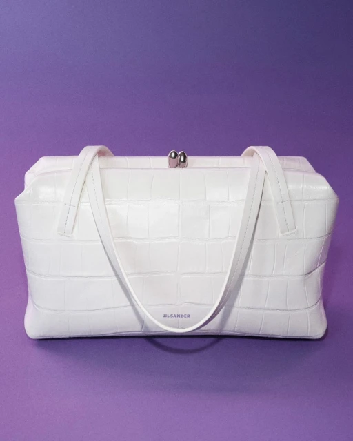 Studio shot of a white Jil Sander handbag with a crocodile print set against a solid purple backdrop.