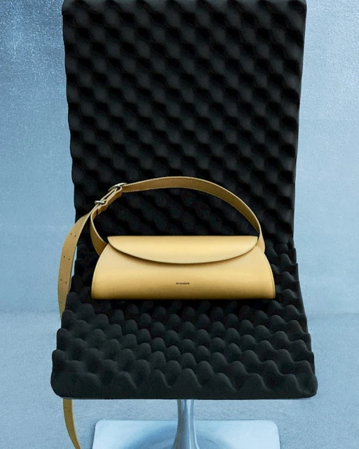 Beige leather handbag with a shoulder strap displayed on a textured black chair against a light blue and gray background.