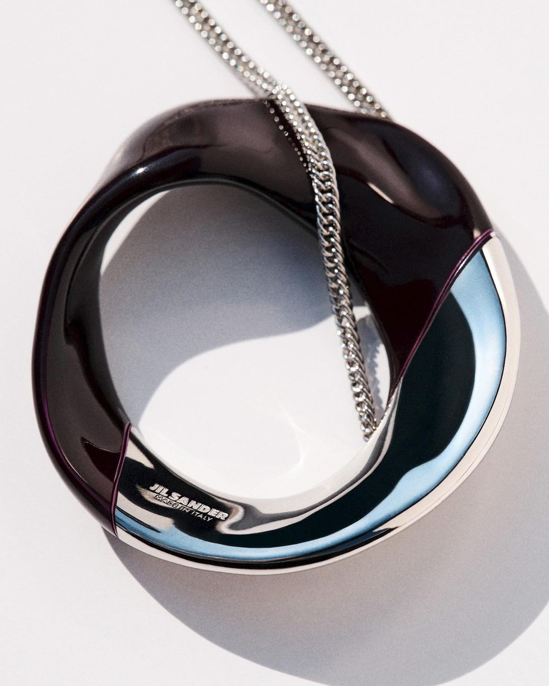 Fashion product shot of a Jil Sander handbag featuring a shiny, dark brown and silver circular body with a delicate silver chain.