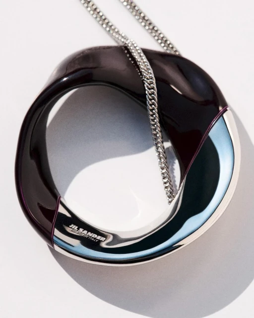 Fashion product shot of a Jil Sander handbag featuring a shiny, dark brown and silver circular body with a delicate silver chain.