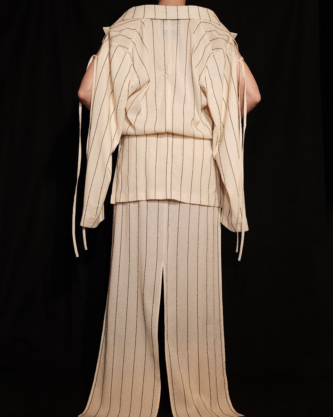 Back view of a person in a cream-colored, vertically striped matching set, including a jacket and pants, against a black backdrop.