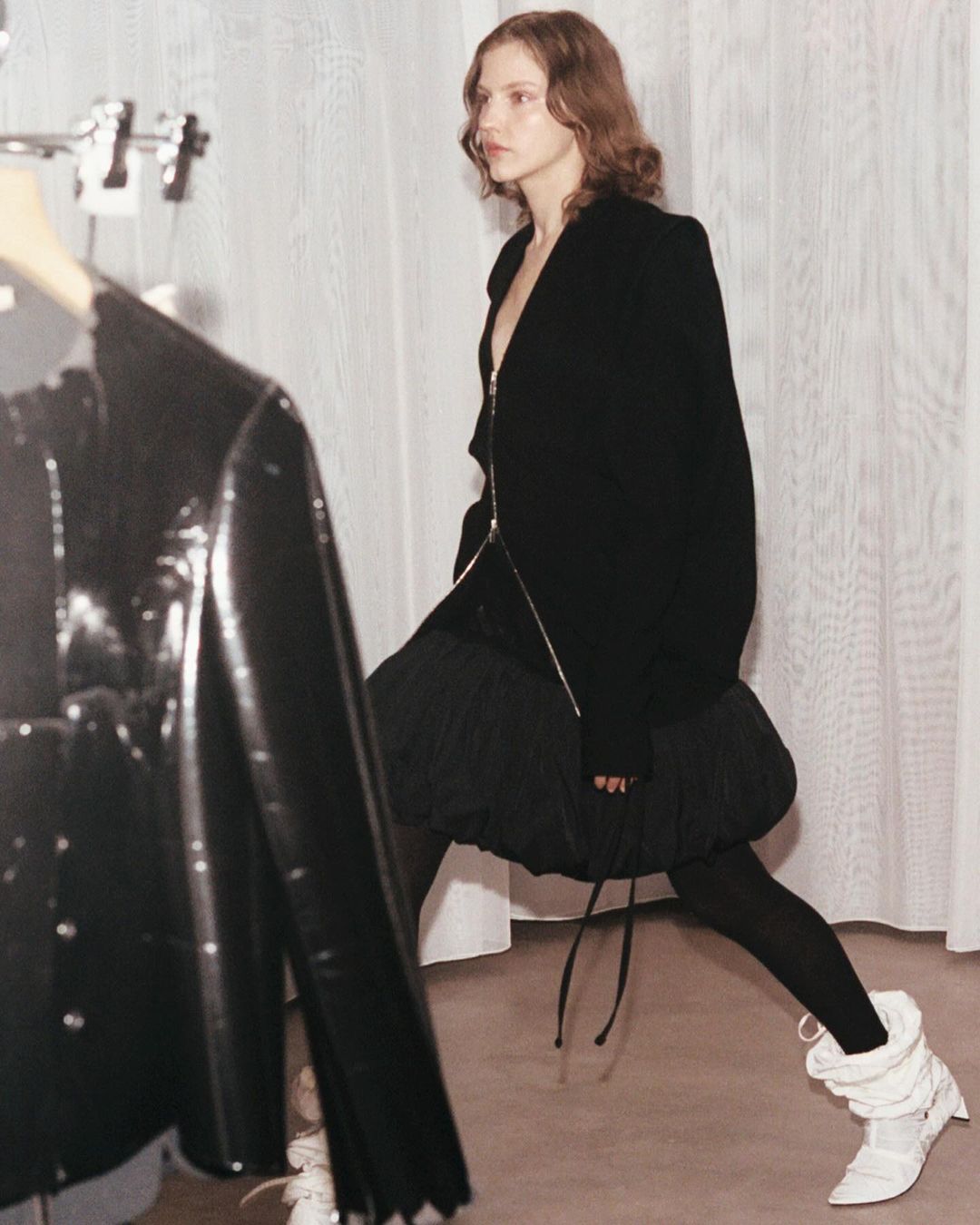 Fashion editorial shot of a tall adult female posing in a black zipper jacket, black skirt, leggings and white boots, in a white indoor space.