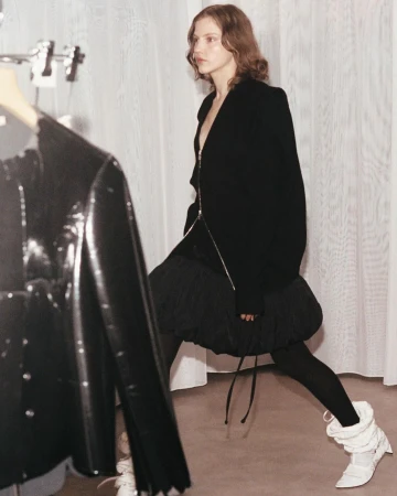 Fashion editorial shot of a tall adult female posing in a black zipper jacket, black skirt, leggings and white boots, in a white indoor space.