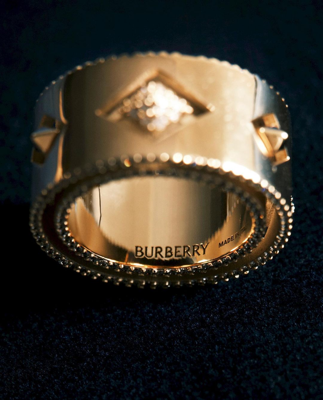 Close-up of a shiny gold ring with geometric pattern on a black surface; the ring is inscribed with 'BURBERRY'.