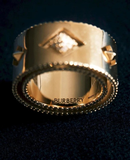 Close-up of a shiny gold ring with geometric pattern on a black surface; the ring is inscribed with 'BURBERRY'.