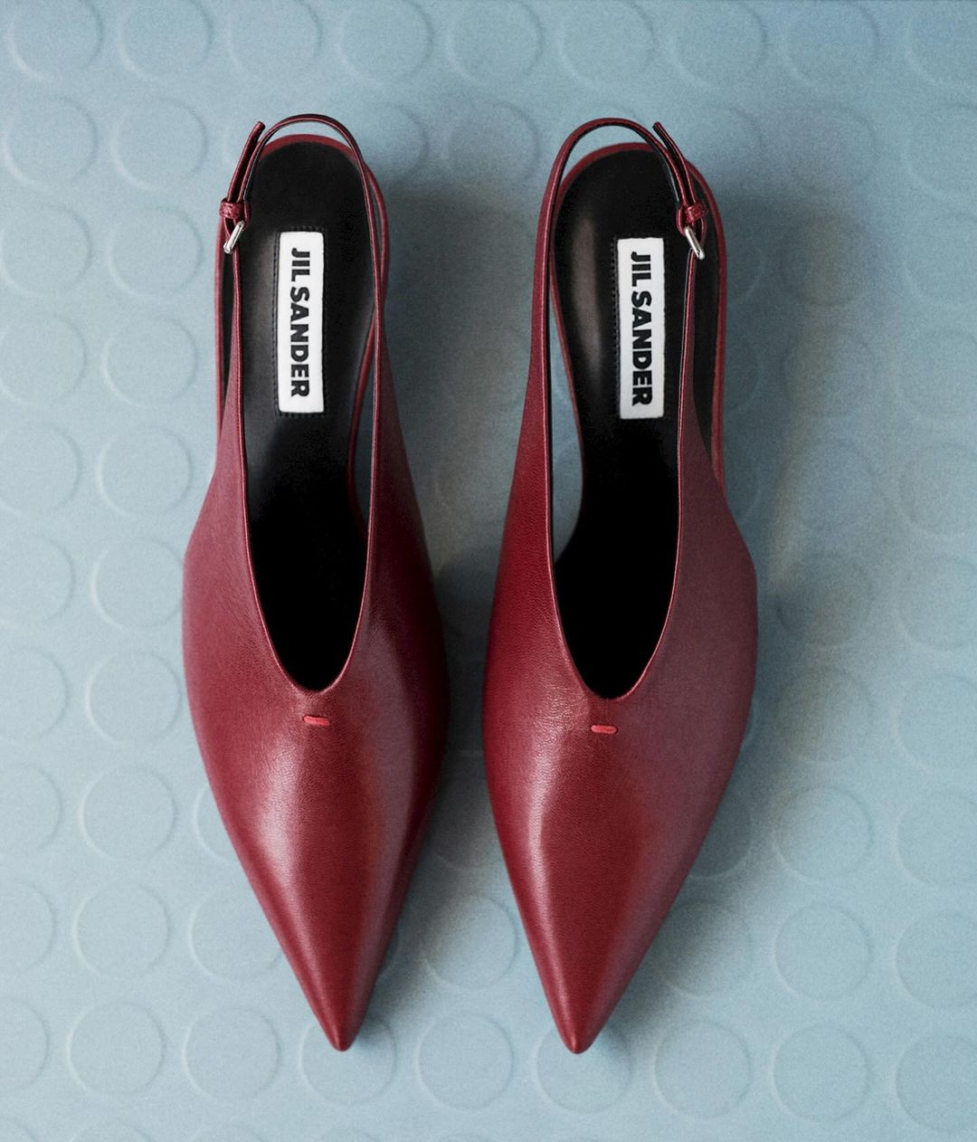 Overhead shot of a pair of red leather Jil Sander slingback heels with a pointed toe on a light blue background with circular pattern.