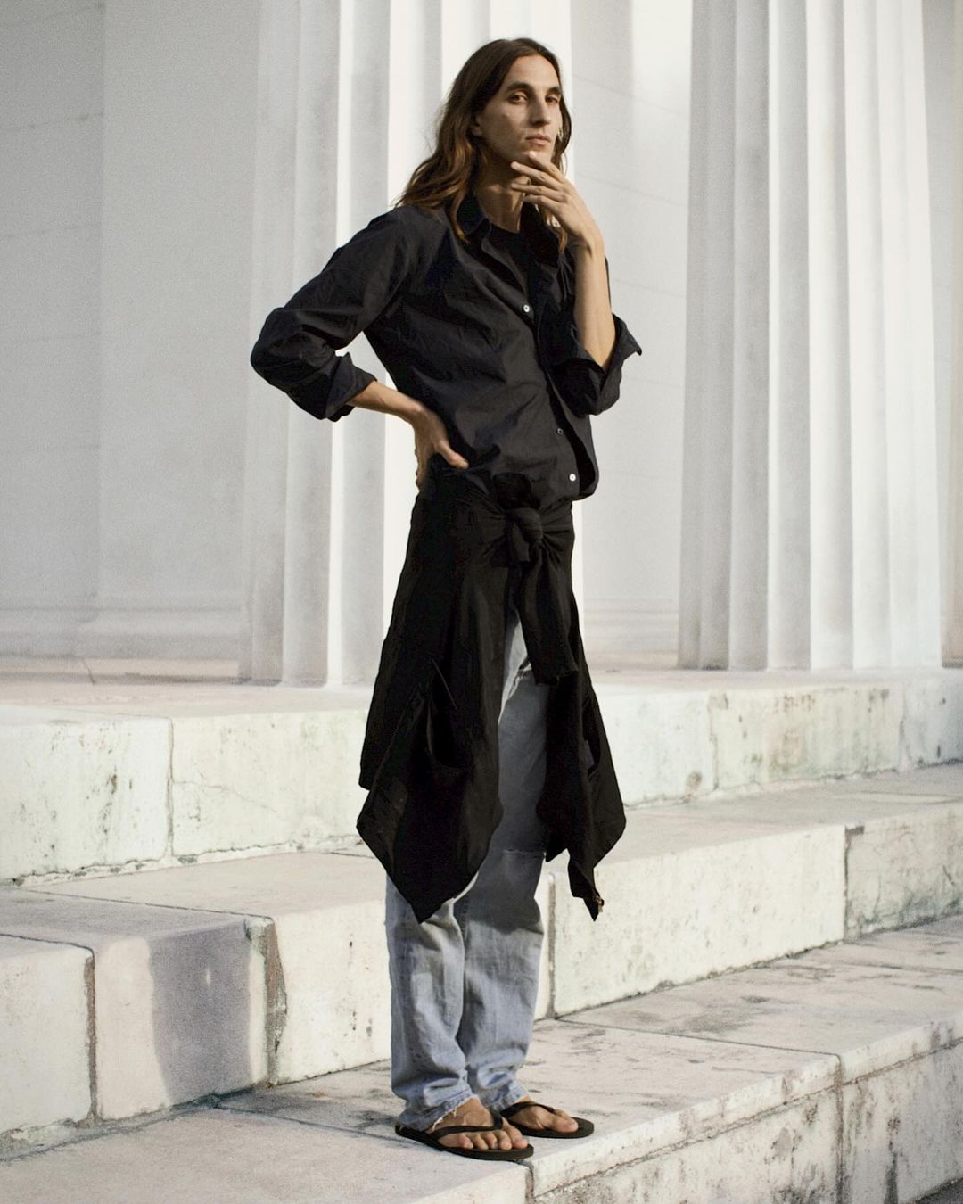 A long-haired woman in a black button-down shirt, grey pants, and sandals poses in front of white columns on stone steps in daylight.