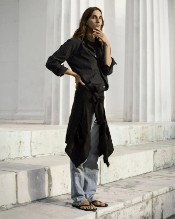 A long-haired woman in a black button-down shirt, grey pants, and sandals poses in front of white columns on stone steps in daylight.