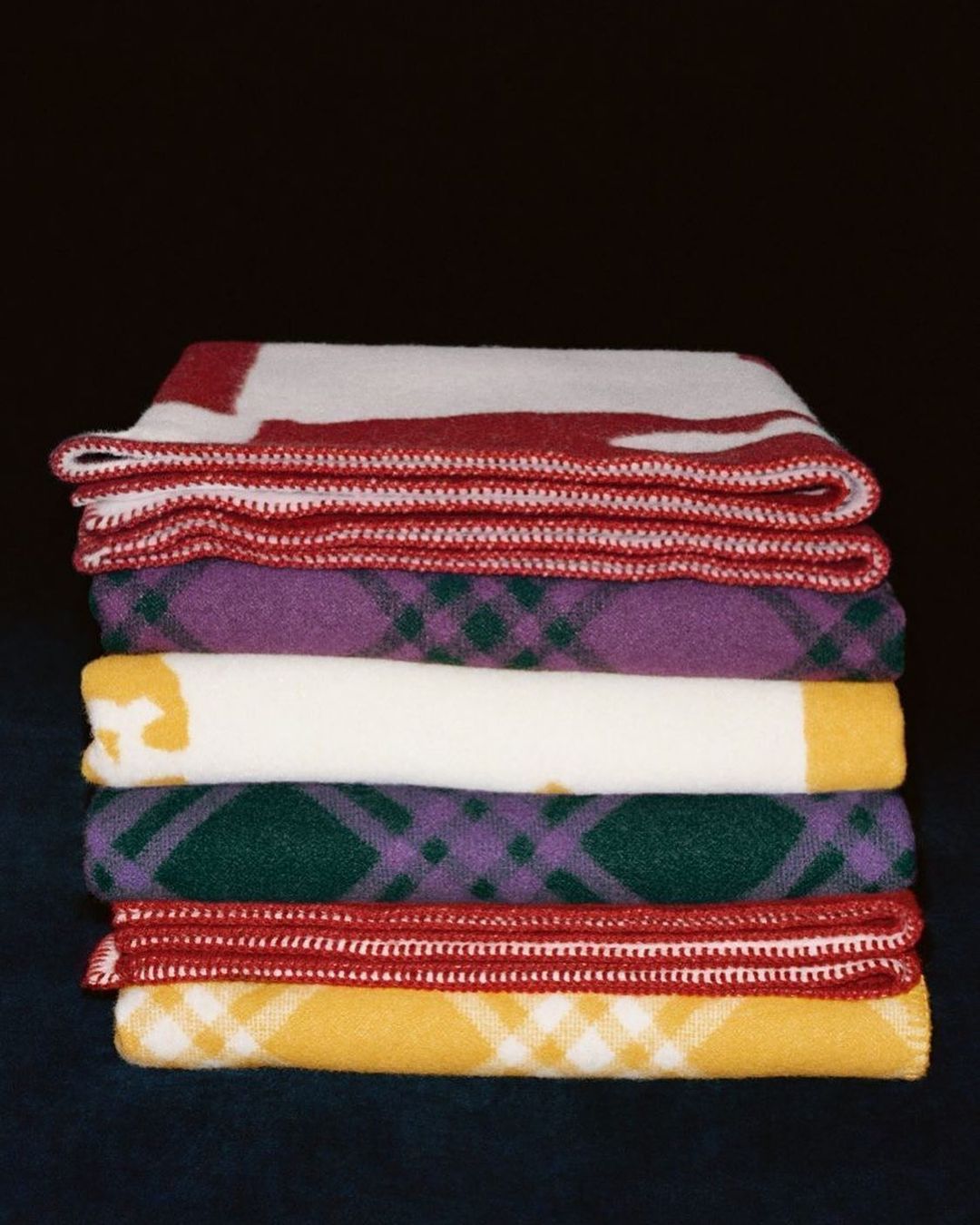 A stack of colorful, patterned wool blankets with red, yellow, green, white, and purple tones against a dark background.