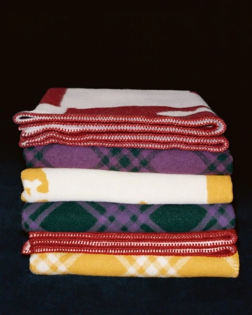 A stack of colorful, patterned wool blankets with red, yellow, green, white, and purple tones against a dark background.