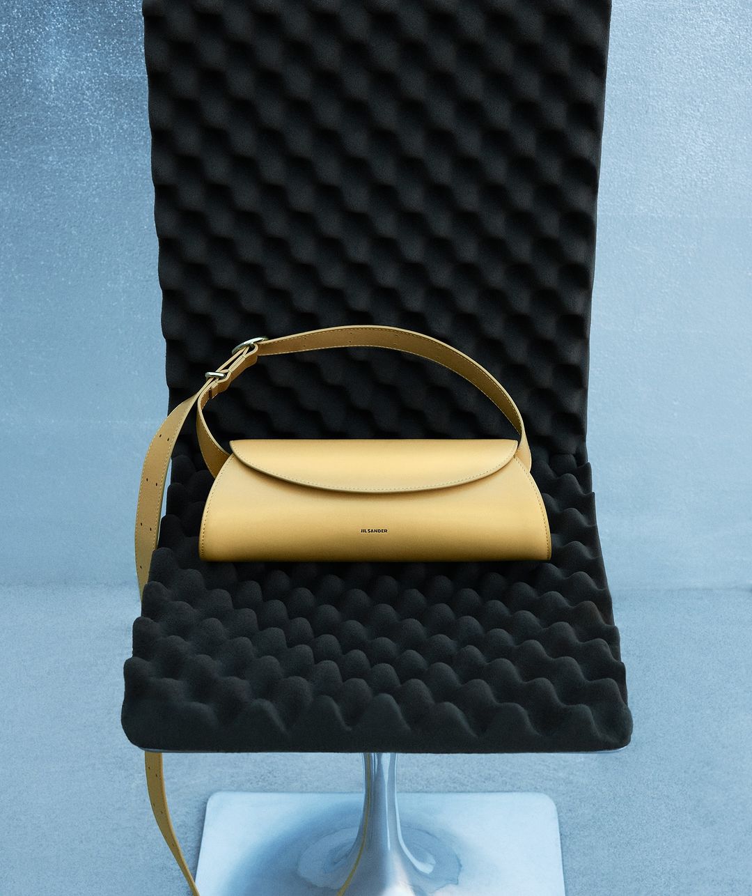 Tan leather handbag with a shoulder strap on a black textured chair against a blue-grey wall in a studio setting, product shot.