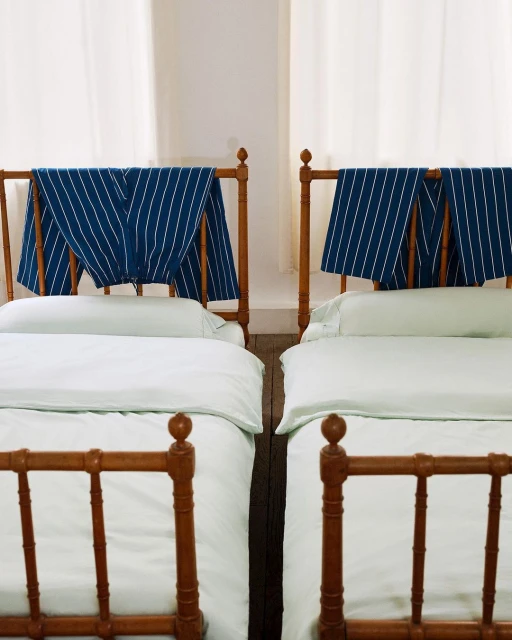 Two beds with white bedding and wooden frames in a bright room, each featuring blue and white striped fabric draped over the headboard.