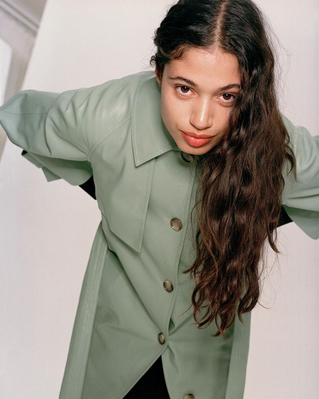Portrait of a brunette woman with long wavy hair wearing a light green trench coat, posing with hands on hips in a bright studio.
