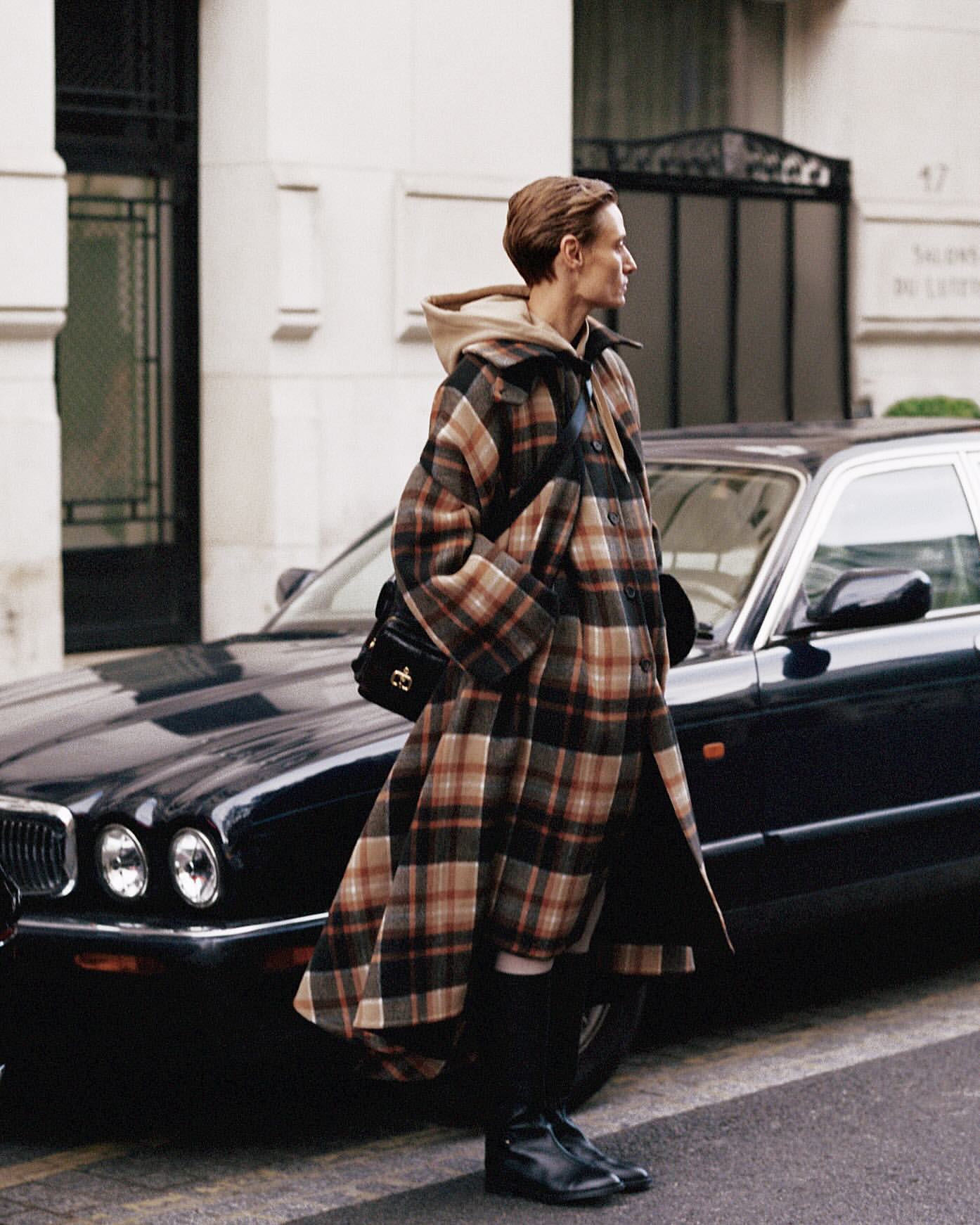 Woman in a plaid coat stands next to a parked black Jaguar car on a city street, with a building visible in the background on a cloudy day.