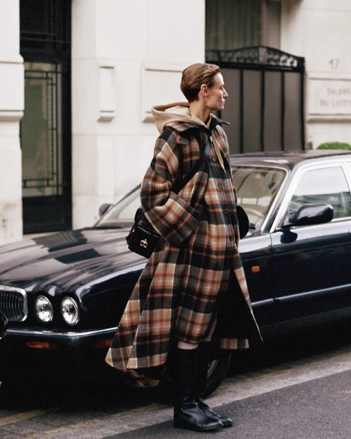 Woman in a plaid coat stands next to a parked black Jaguar car on a city street, with a building visible in the background on a cloudy day.