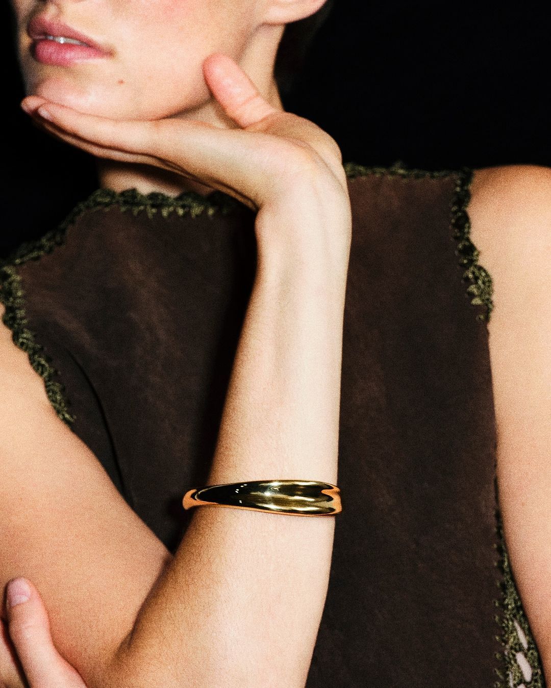 A close-up shot showcases a woman's arm adorned with a gold bangle bracelet, with her hand near her chin, set against a dark backdrop.