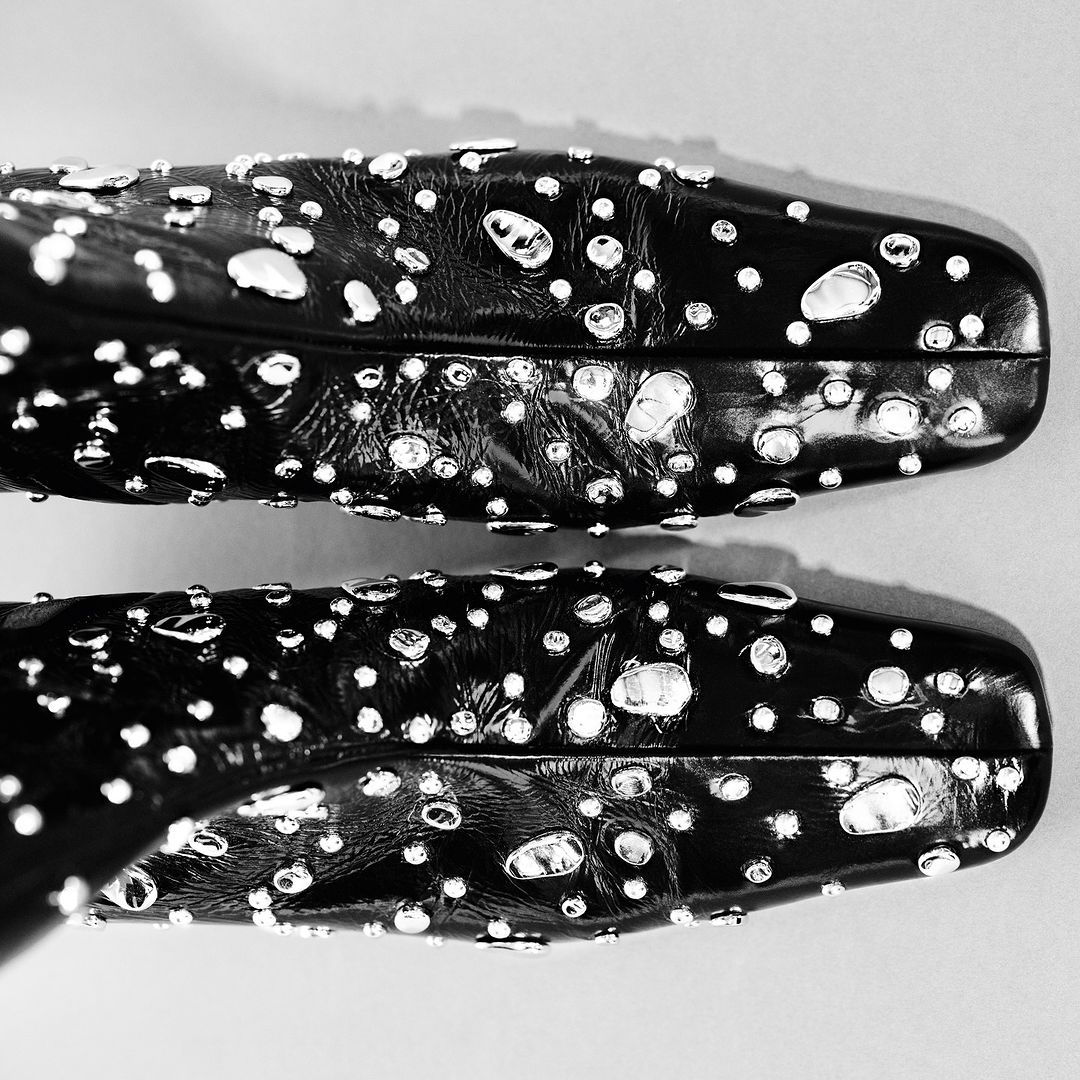 Top-down studio shot of two black patent leather heeled shoes adorned with silver studs, against a light gray seamless backdrop.