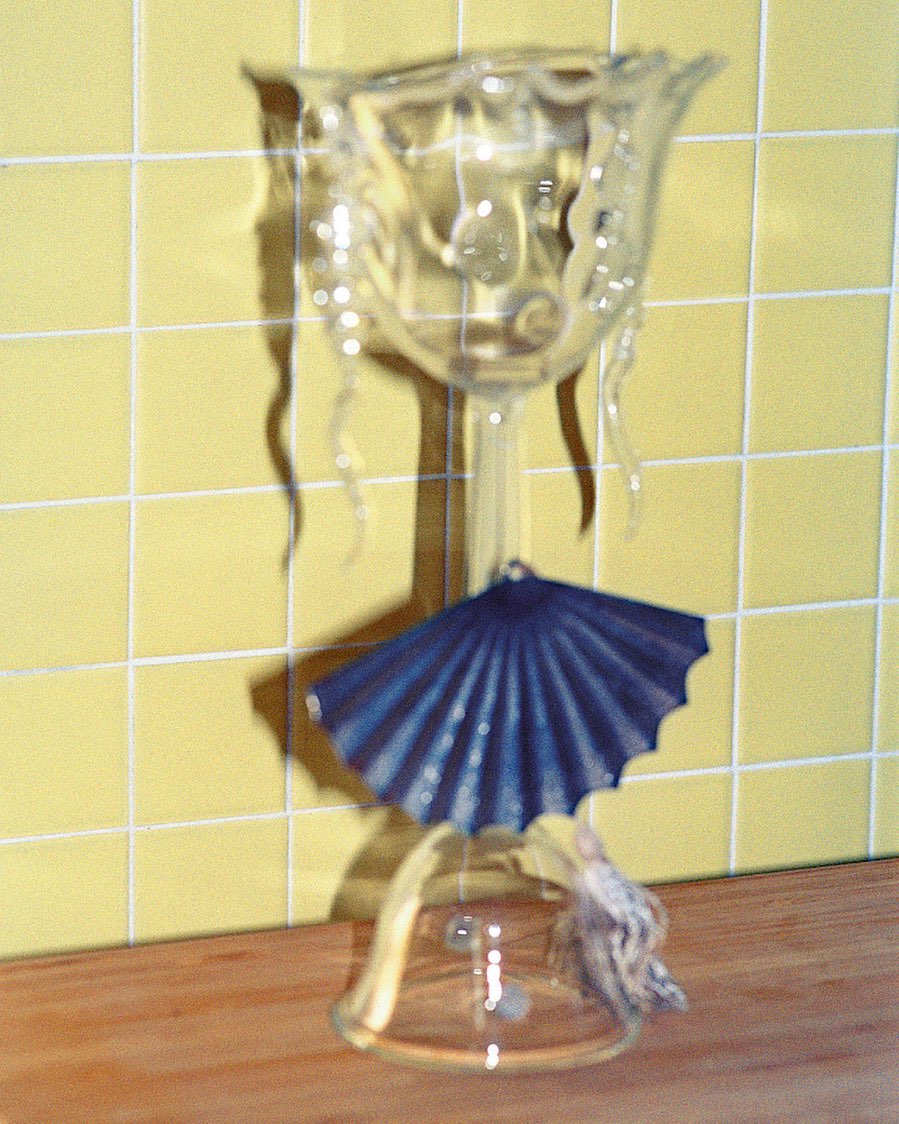 Clear glass decorative chalice with a small dark blue paper fan hanging from it sits on a wooden surface in front of a yellow tiled wall.