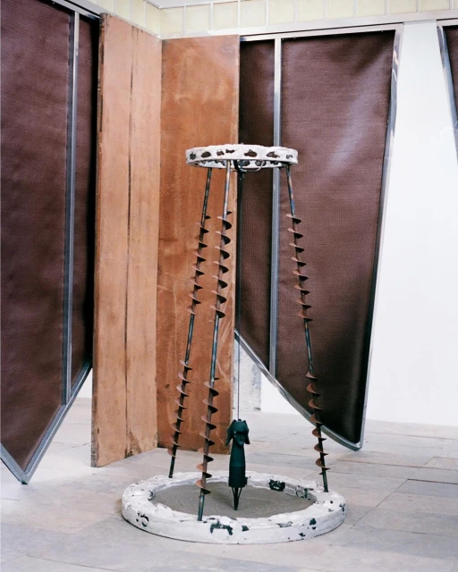 Abstract photo of sculptural artwork featuring metal rods, wooden disks, and a small figure, set against geometric background panels.